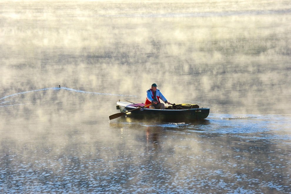 Рыбак плывет на лодке сквозь пелену утреннего тумана на озере Титизее в Шварцвальде, Германия.